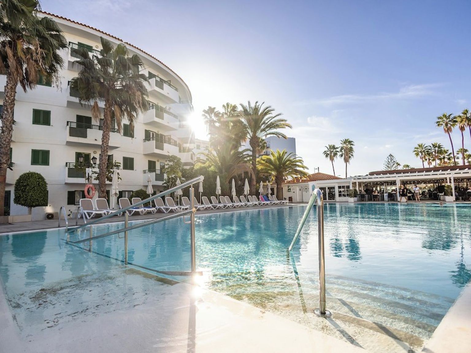 Utomhuspool på en solig resort med palmer och en vit byggnad.