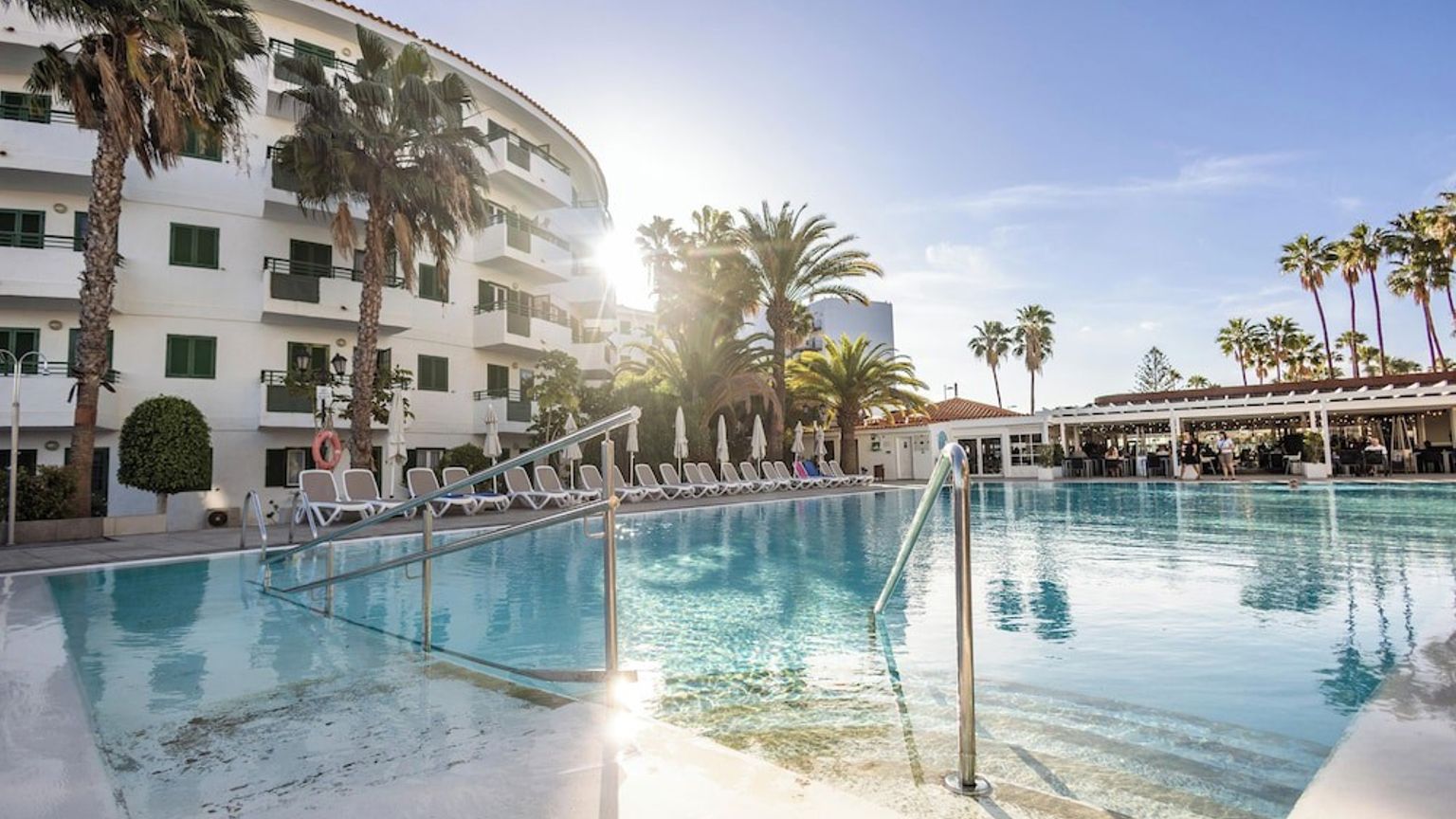Utomhuspool på en solig resort med palmer och en vit byggnad.