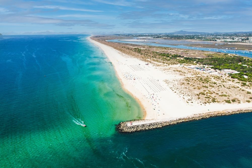 Flygfoto över en lång sandstrand med klart turkost vatten, en stenpir och rader av solstolar.