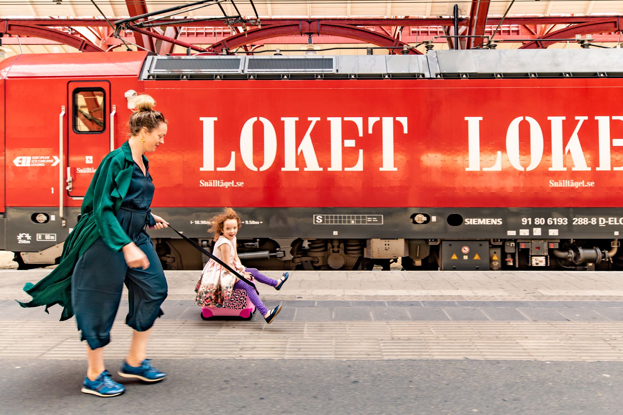 En kvinna drar ett leende barn som sitter på en rosa resväska på en tågplattform med ett rött tåg i bakgrunden.