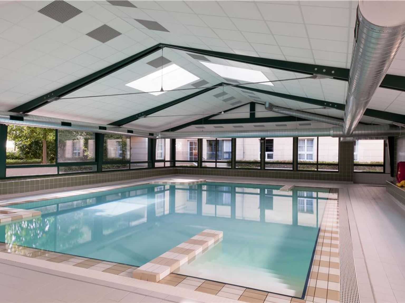 Et innendørs svømmebasseng med klart blått vann under et høyt, skrått tak med takvinduer og store vinduer. Adagio Aparthotel Val d'Europe nær Disneyland® Paris