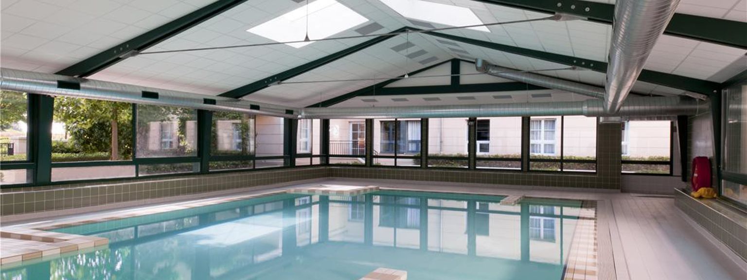 Et innendørs svømmebasseng med klart blått vann under et høyt, skrått tak med takvinduer og store vinduer. Adagio Aparthotel Val d'Europe nær Disneyland® Paris