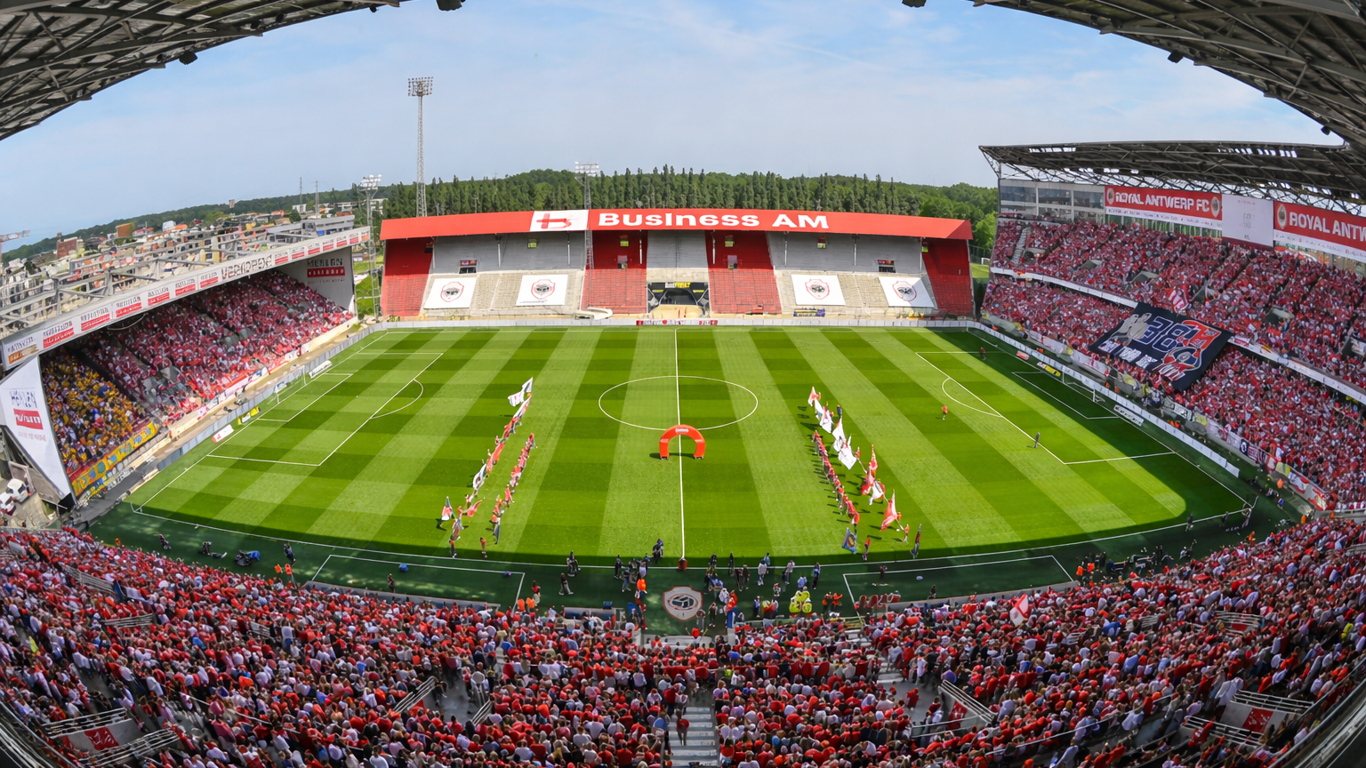 A packed football stadium with a green field, red-clad fans, and "Business AM" signage on the stands.