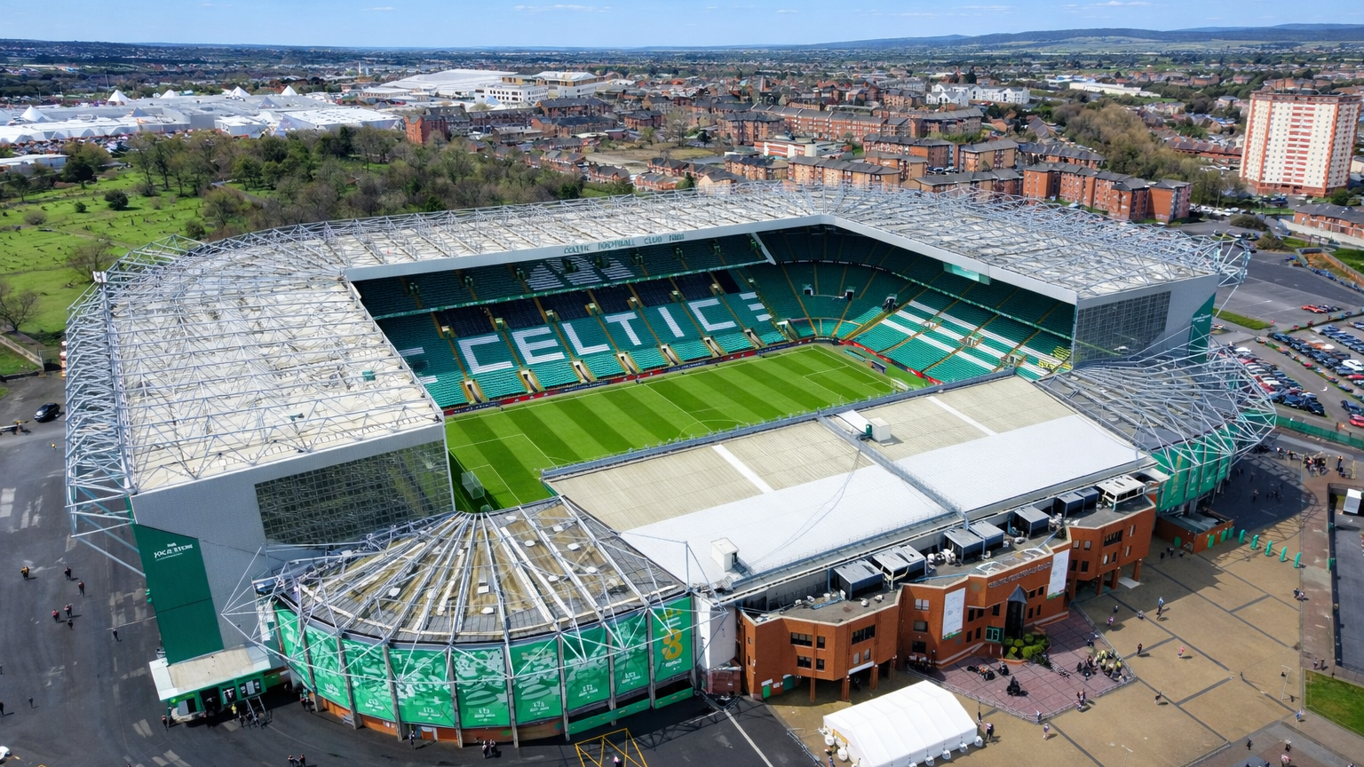 Aerial view of Celtic Park, a large green and white football stadium, surrounded by an urban landscape under a blue sky.
