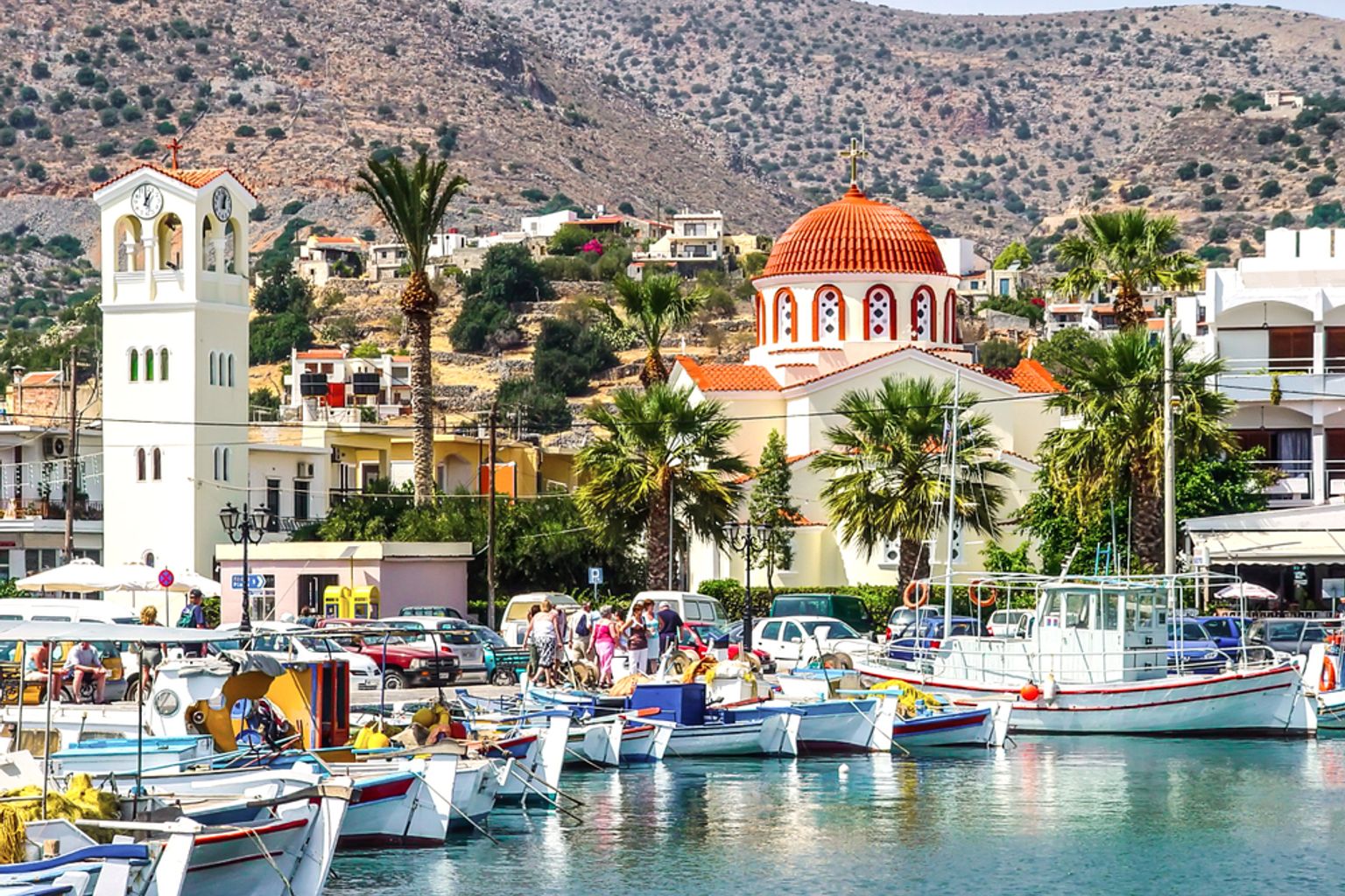 A scenic harbor with many small fishing boats, a white bell tower, and a red-domed church along the shore, backed by hills. Elounda, Greece
