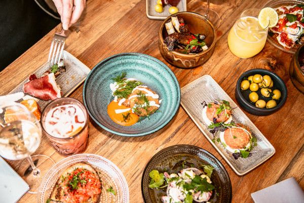 Udsigt oppefra af et træbord fyldt med adskillige tallerkener med forretter, drikkevarer og en hånd, der holder en gaffel.