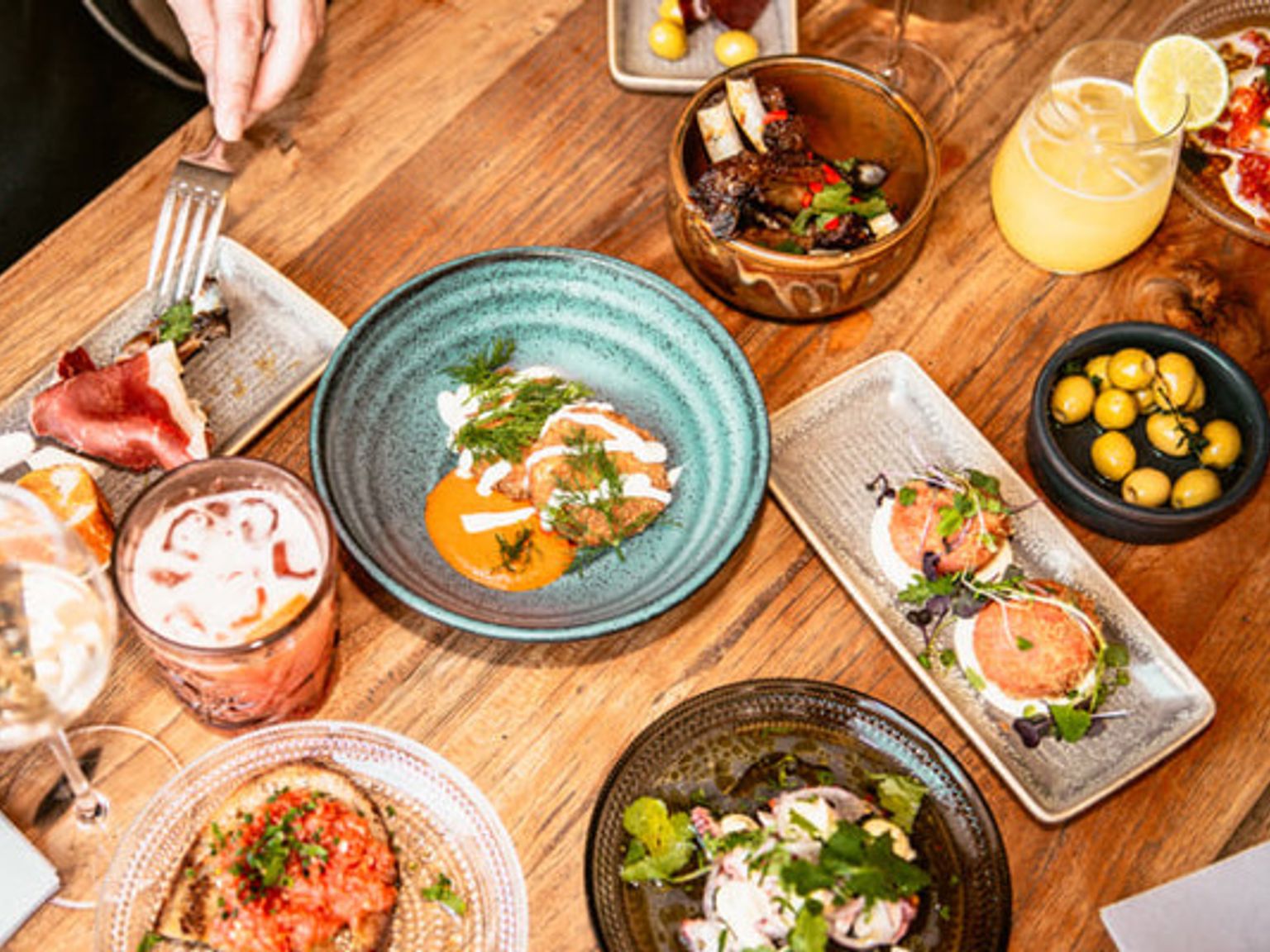 Udsigt oppefra af et træbord fyldt med adskillige tallerkener med forretter, drikkevarer og en hånd, der holder en gaffel.