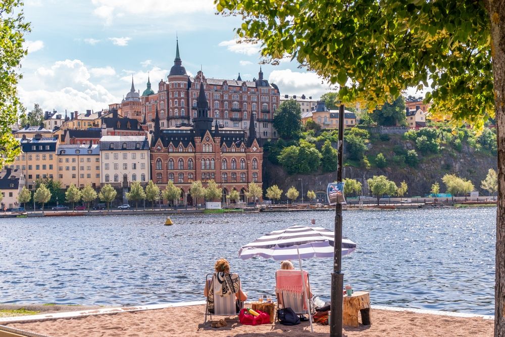 To personer slapper af under en parasol på en sandstrand, vendt mod en by med historiske bygninger på den anden side af vandet.