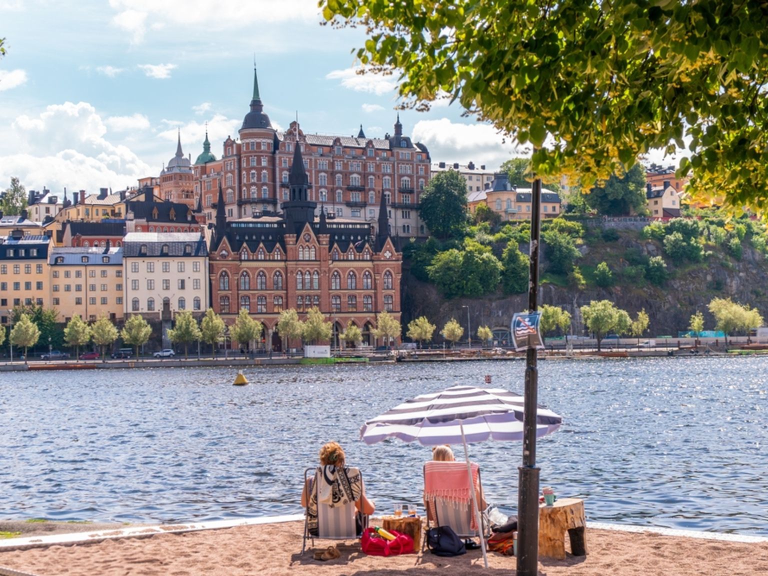 To personer slapper af under en parasol på en sandstrand, vendt mod en by med historiske bygninger på den anden side af vandet.