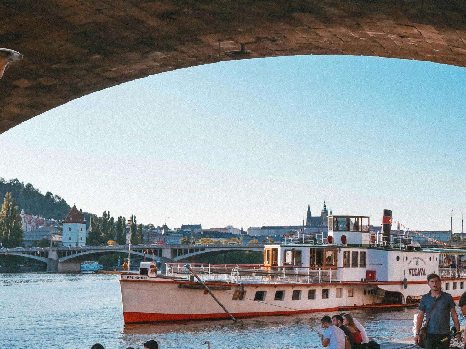 en grupp människor sitter vid stranden av en flod med en båt i bakgrunden som säger pizza