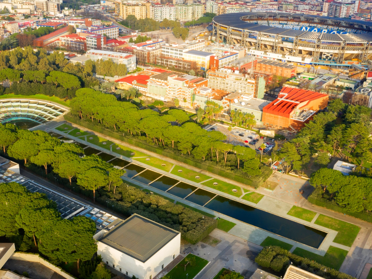 Aerial view of a city park with water features and a unique curved building, next to a large stadium showing 'NAPOLI'.