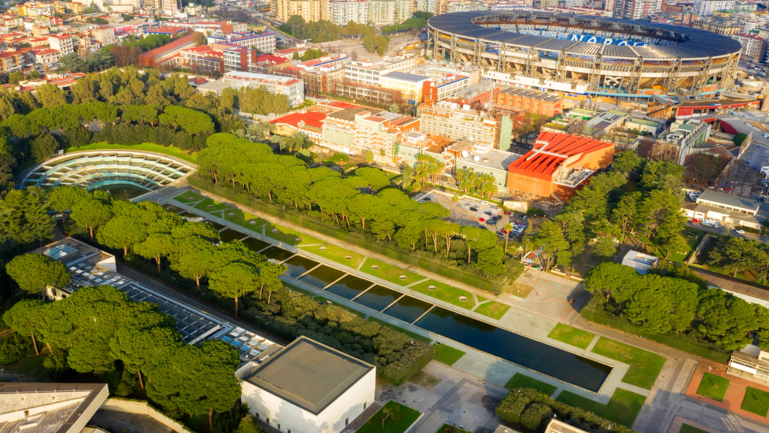 Aerial view of a city park with water features and a unique curved building, next to a large stadium showing 'NAPOLI'.