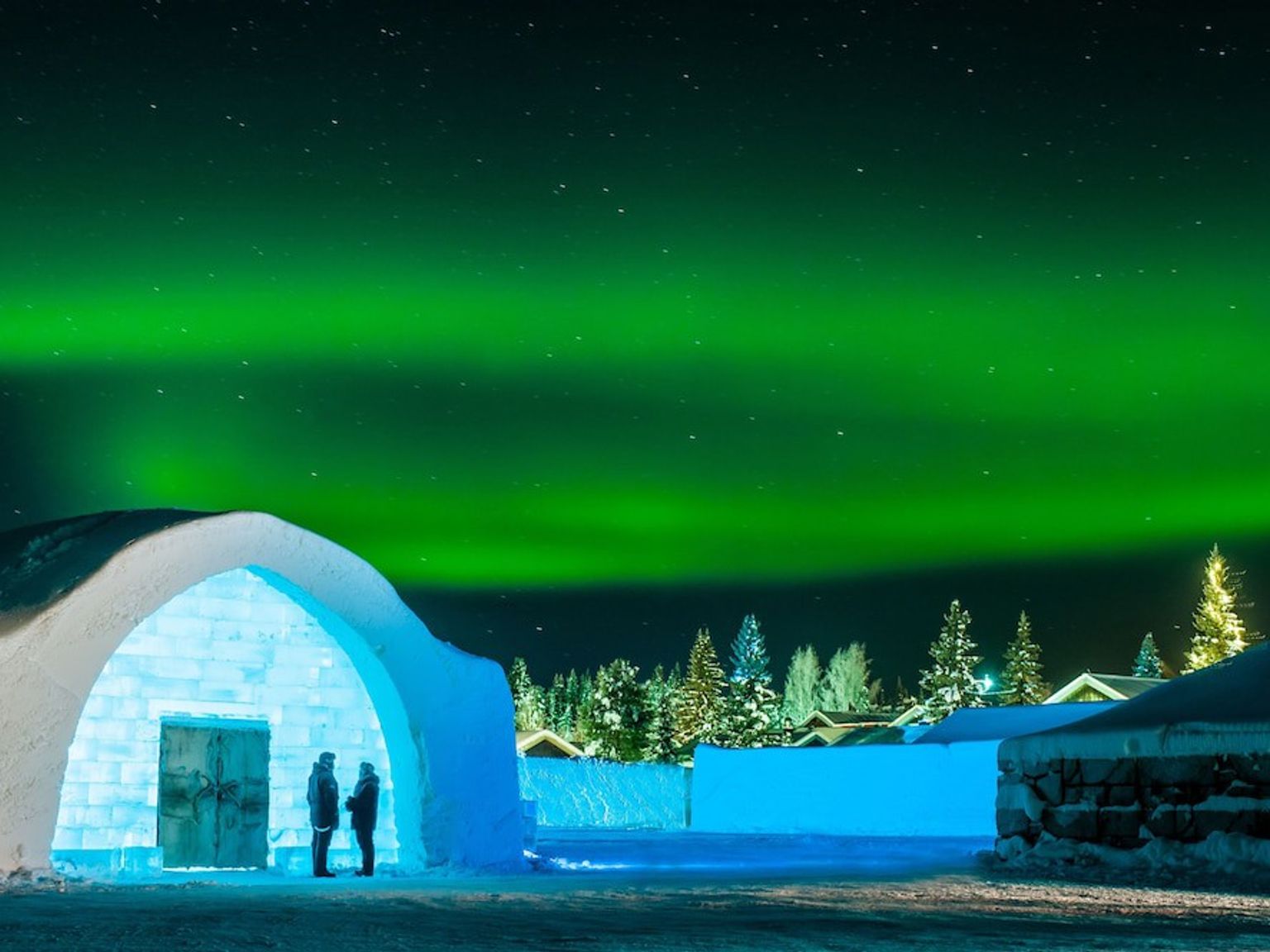 dos personas están de pie frente a una casa de hielo bajo la aurora boreal