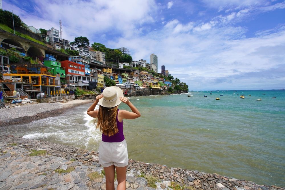Bagfra af en kvinde med stråhat, der kigger på en farverig bjergskråningsby ved havet med både.