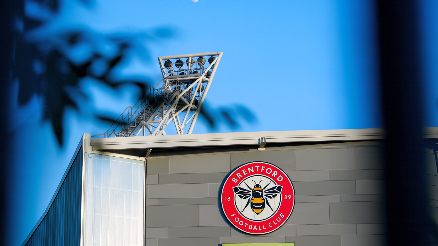 Brentford Football Club stadium building with its logo, a floodlight, and the moon against a blue sky.