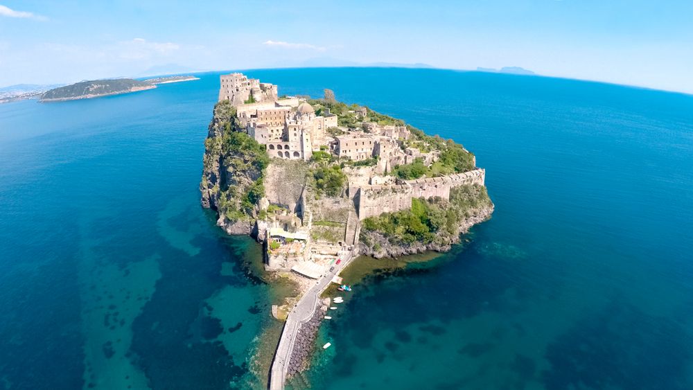 Ancienne forteresse sur une île rocheuse reliée par une chaussée, entourée d’une mer bleu clair. Ischia, Italie