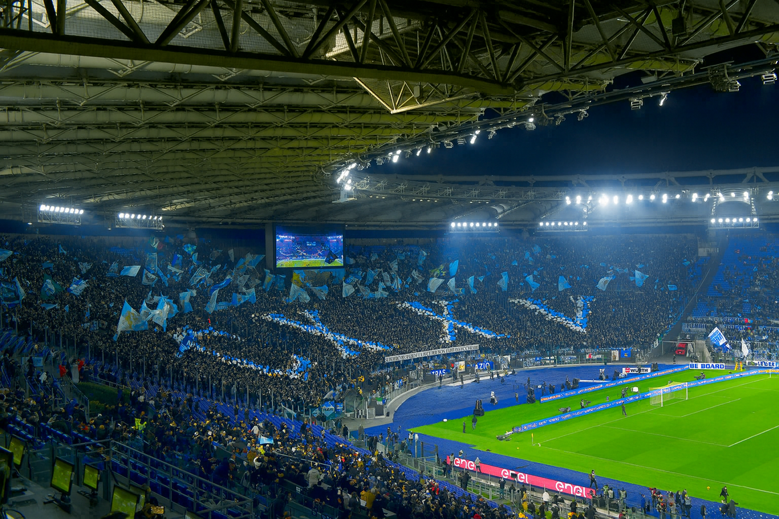 Estadio de fútbol lleno por la noche con aficionados sosteniendo banderas azules para formar un gran patrón de cruz en las gradas.