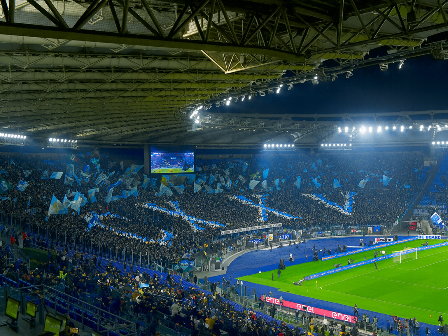 Fullsatt fotbollsstadion på natten med fans som håller blå flaggor för att bilda ett stort korsmönster på läktarna.