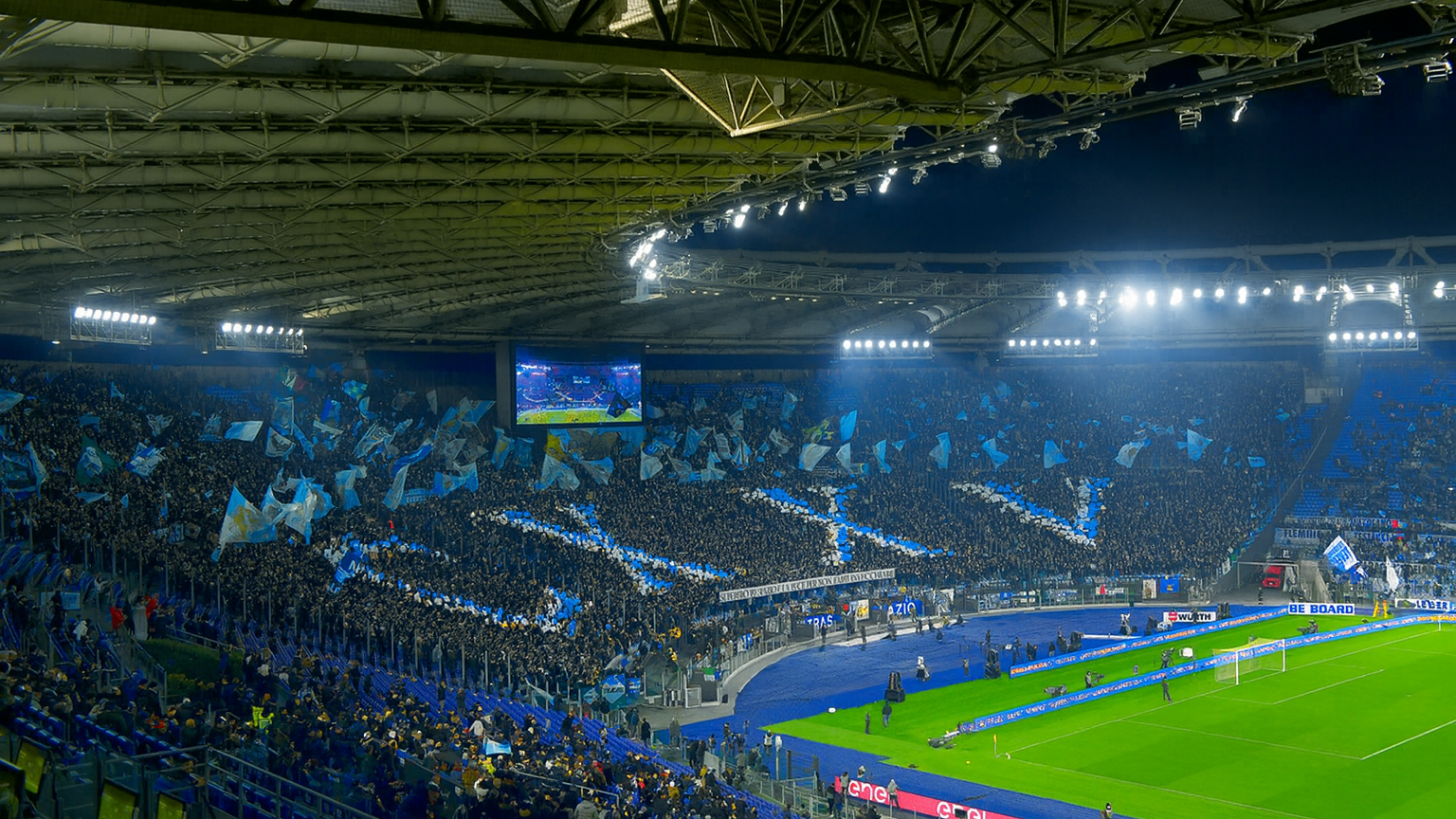 Estadio de fútbol lleno por la noche con aficionados sosteniendo banderas azules para formar un gran patrón de cruz en las gradas.