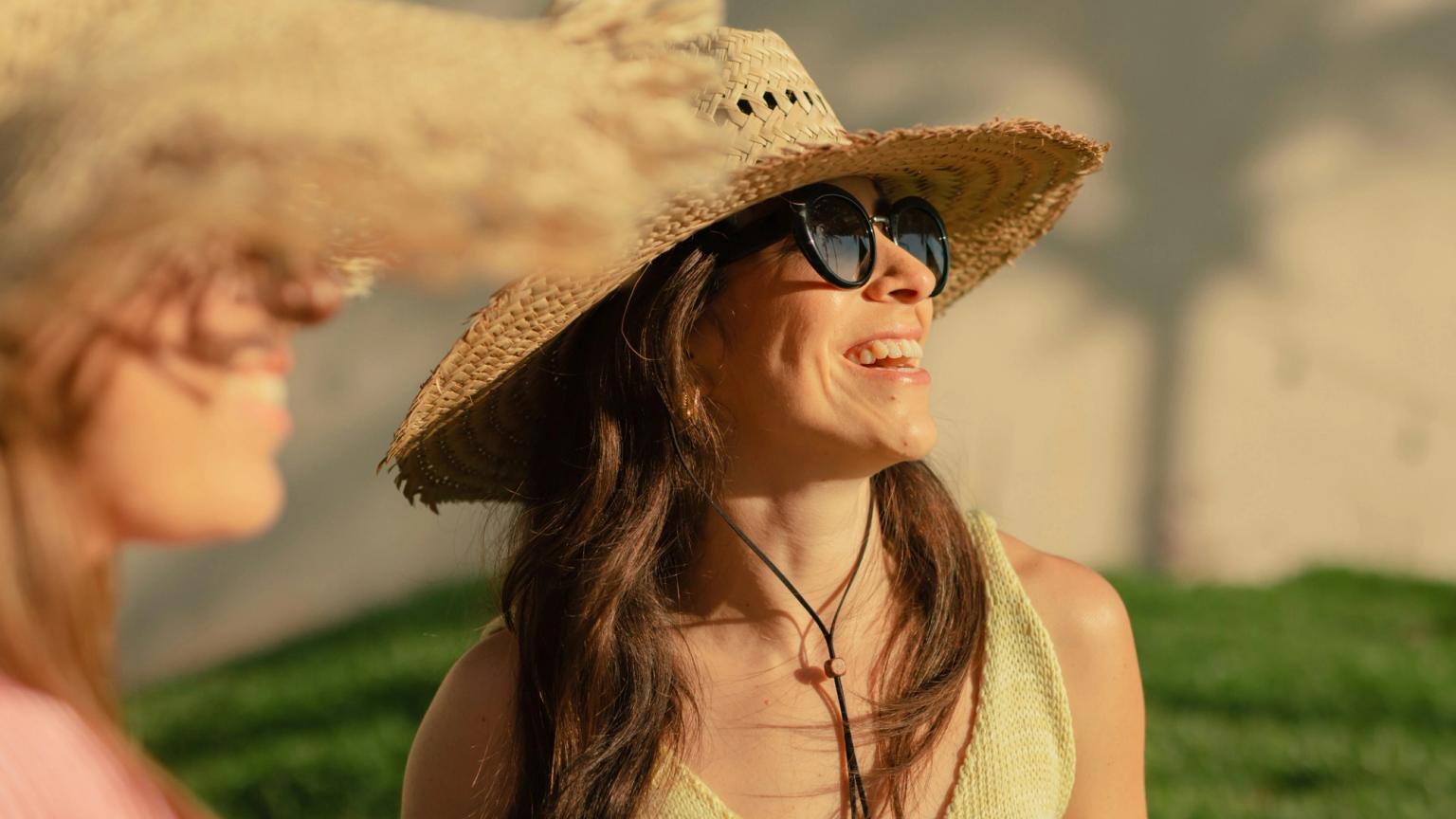 En leende kvinna i en stråhatt och solglasögon tittar upp, med en annan kvinna i en hatt delvis synlig.