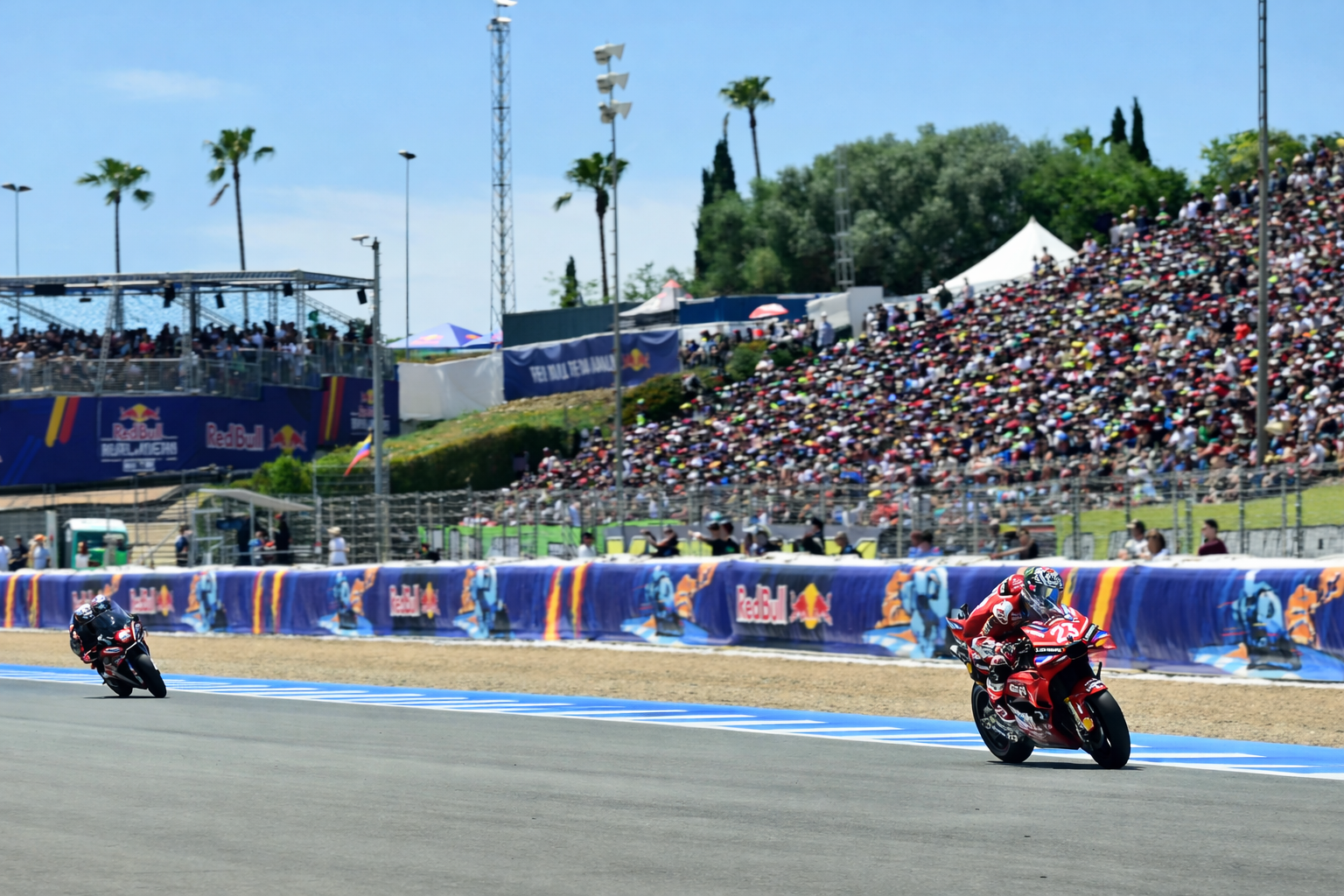 Två motorcykelracingförare lutar sig in i en kurva på en bana, med en stor publik och sponsorbanderoller i bakgrunden.