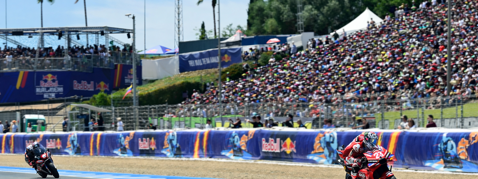 Två motorcykelracingförare lutar sig in i en kurva på en bana, med en stor publik och sponsorbanderoller i bakgrunden.