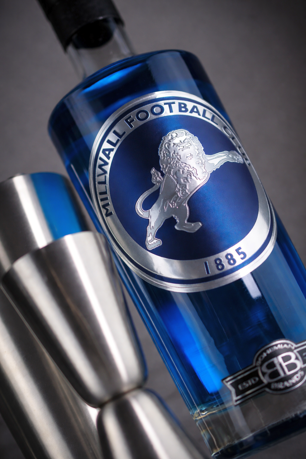 Garrafa azul de bebida Millwall FC com logotipo de leão prateado, ao lado de um medidor de aço inoxidável.