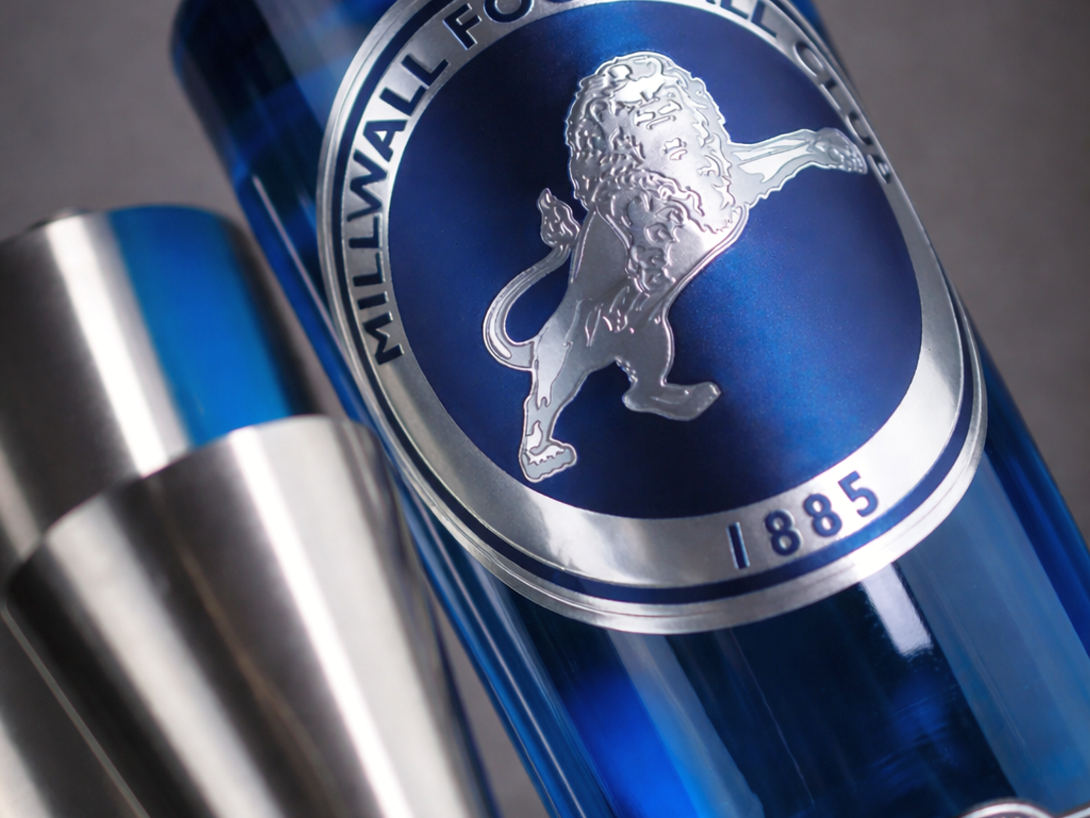 Garrafa azul de bebida Millwall FC com logotipo de leão prateado, ao lado de um medidor de aço inoxidável.