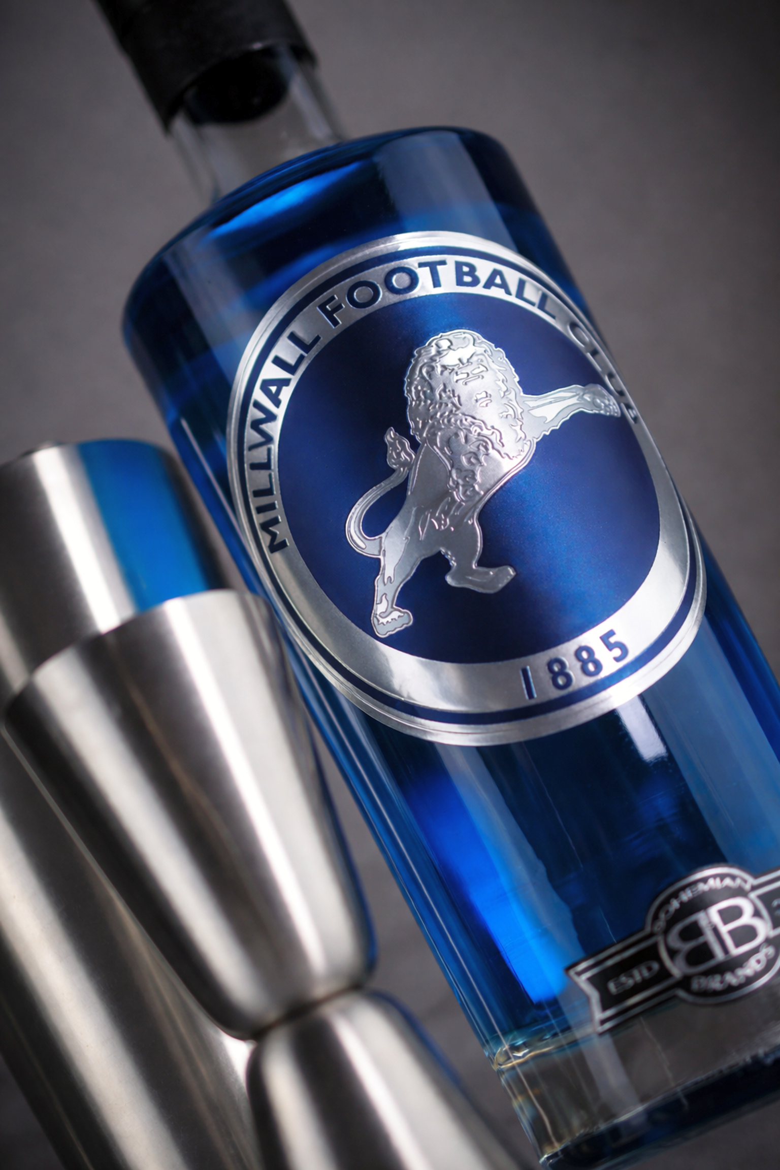 Garrafa azul de bebida Millwall FC com logotipo de leão prateado, ao lado de um medidor de aço inoxidável.