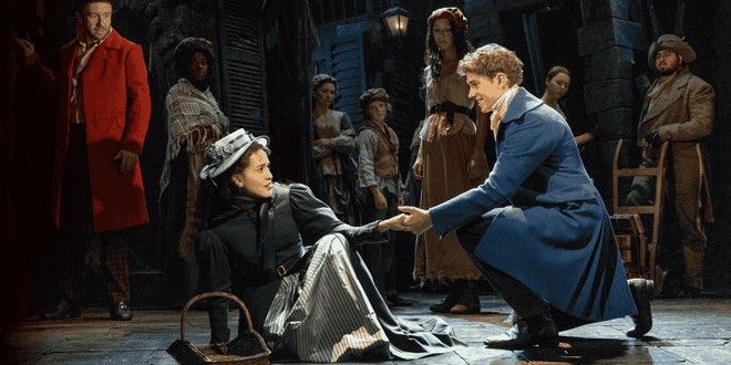 A man in a blue coat kneels, holding the hand of a woman in a dark dress and hat sitting on the ground, surrounded by other characters in period attire on a stage.