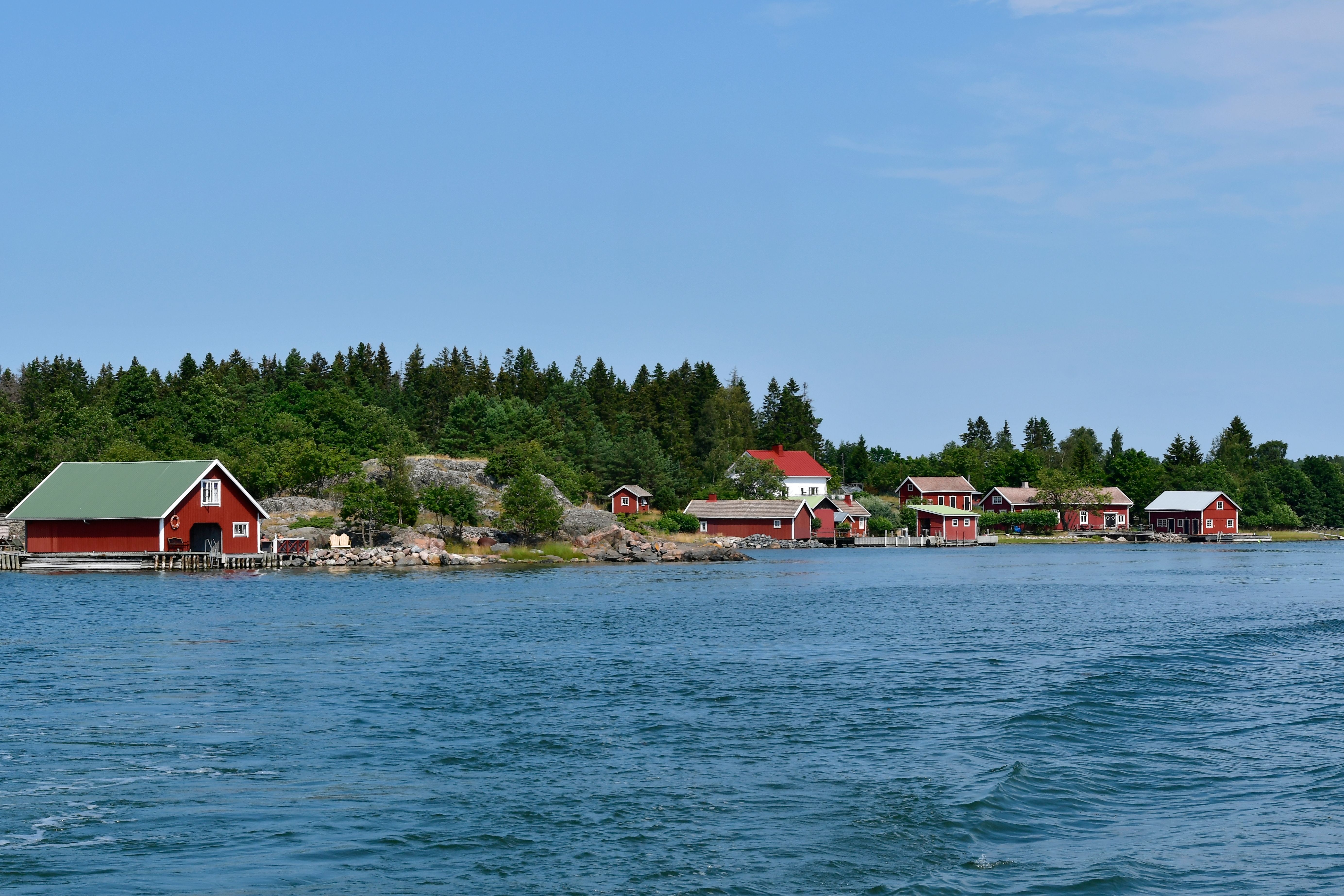 Røde bådehuse og hytter på en klippefyldt, skovklædt kystlinje ved siden af blåt vand.