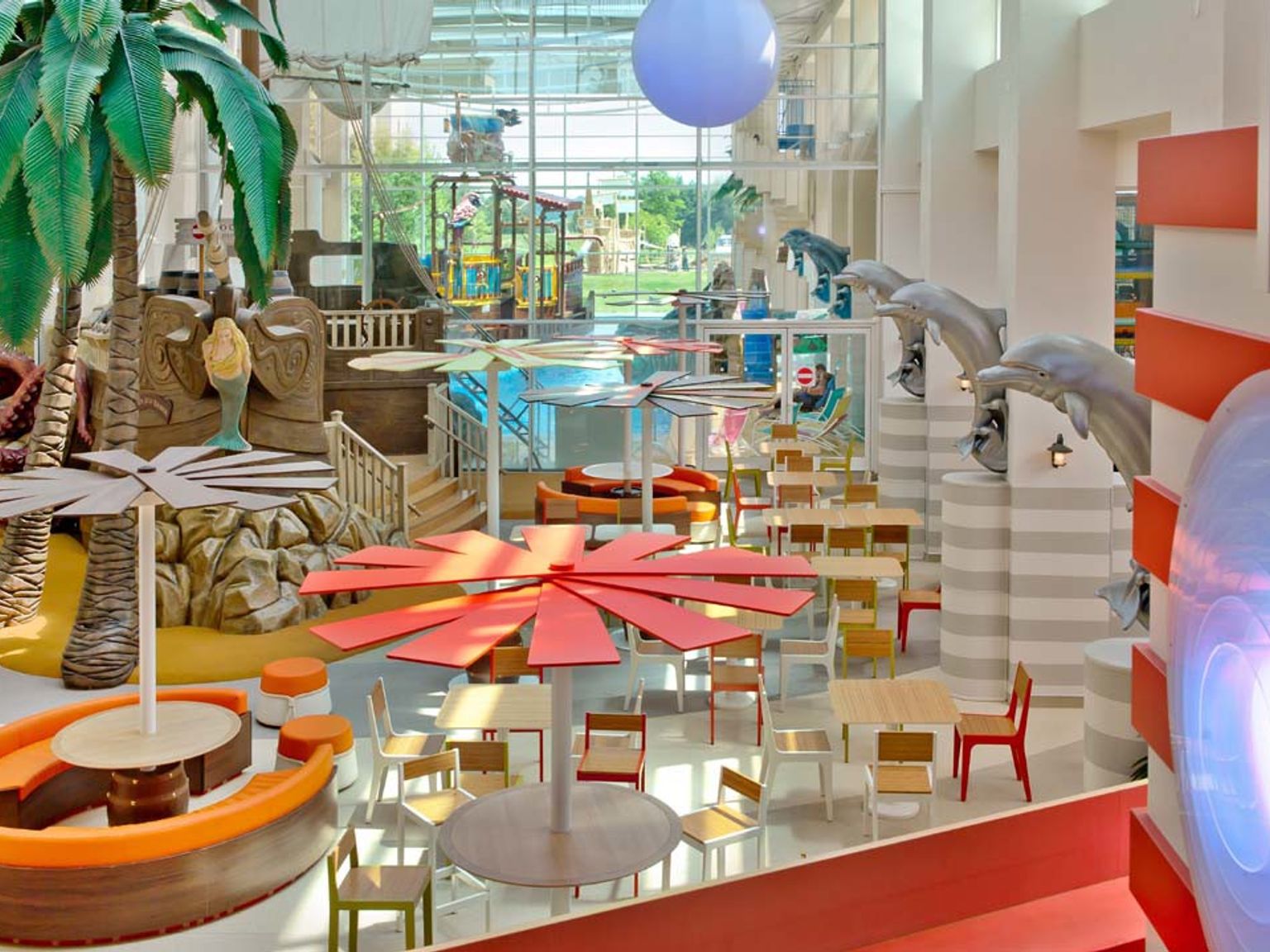 Livlig inomhusmatsal med tropisk inredning, med palmer, pirat-tema lekstrukturer, färgglada bord och delfinskulpturer. Explorers Hotel på Disneyland® Paris