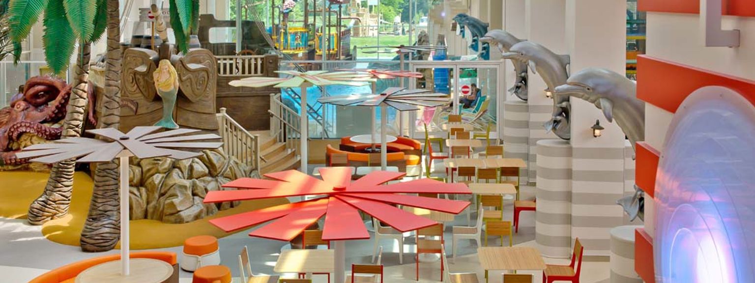 Livlig inomhusmatsal med tropisk inredning, med palmer, pirat-tema lekstrukturer, färgglada bord och delfinskulpturer. Explorers Hotel på Disneyland® Paris