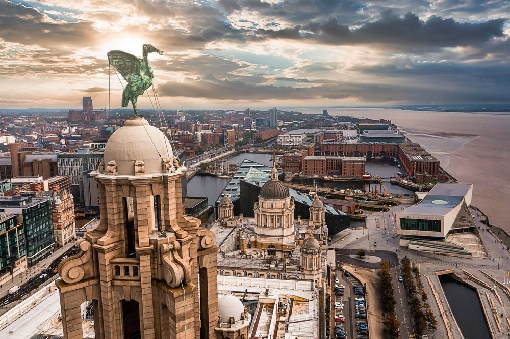 Aerial view of Liverpool showing the Liver Bird statue atop the Liver Building, city skyline, docks, and River Mersey under a dramatic sky.