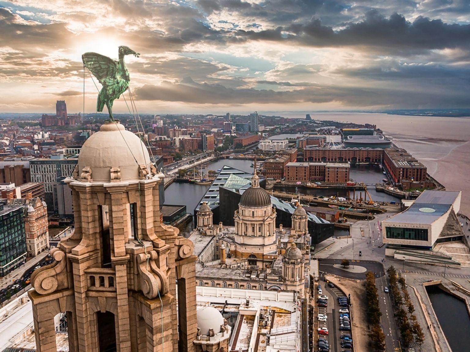 Aerial view of Liverpool showing the Liver Bird statue atop the Liver Building, city skyline, docks, and River Mersey under a dramatic sky.
