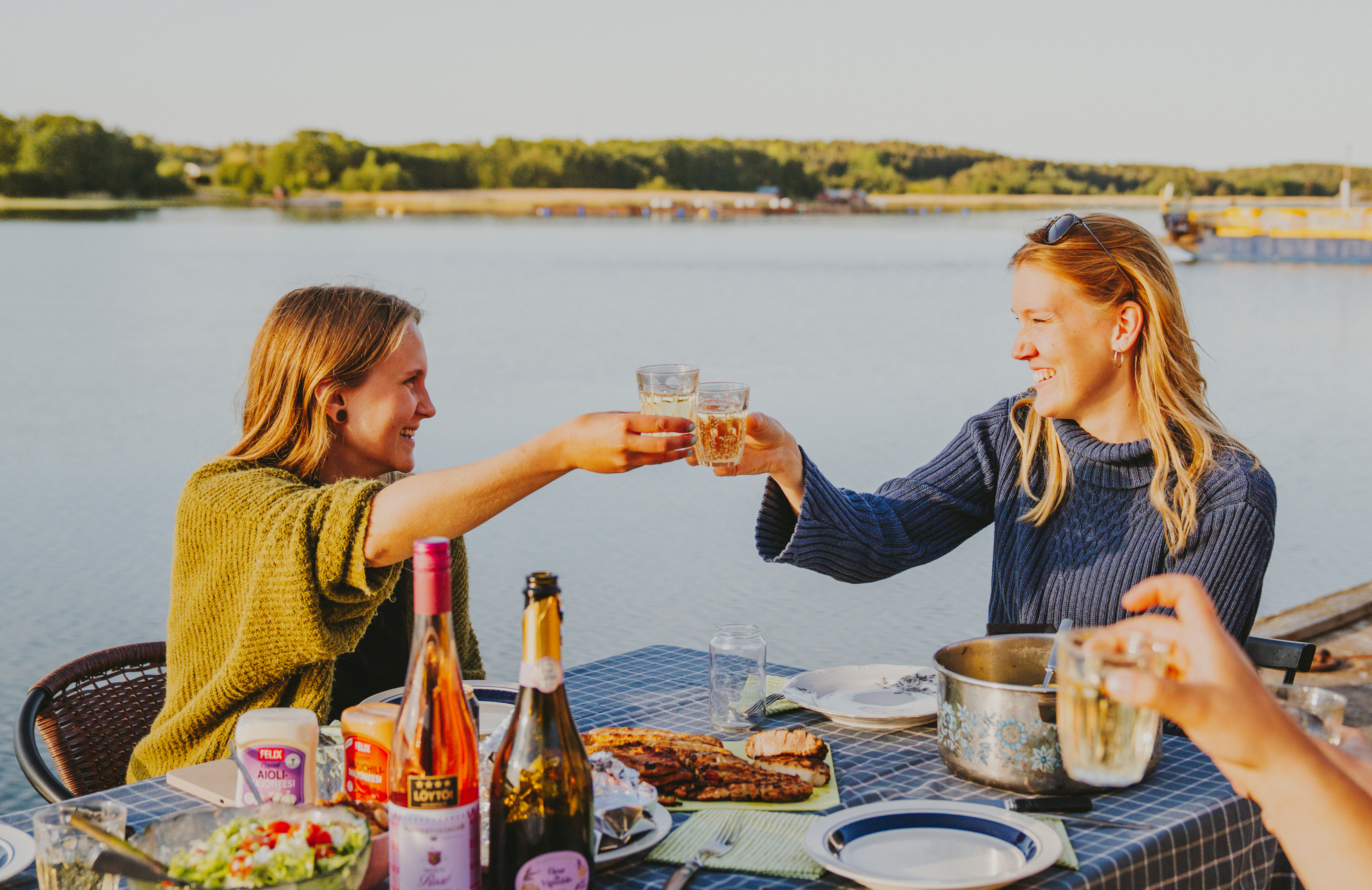To kvinder skåler med drinks ved et udendørs bord fyldt med mad, ved siden af en sø.