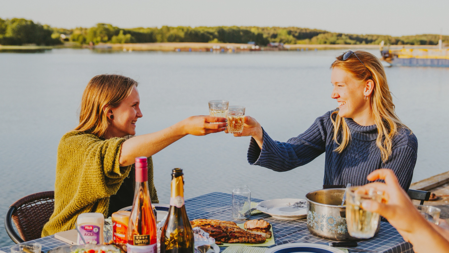 To kvinder skåler med drinks ved et udendørs bord fyldt med mad, ved siden af en sø.