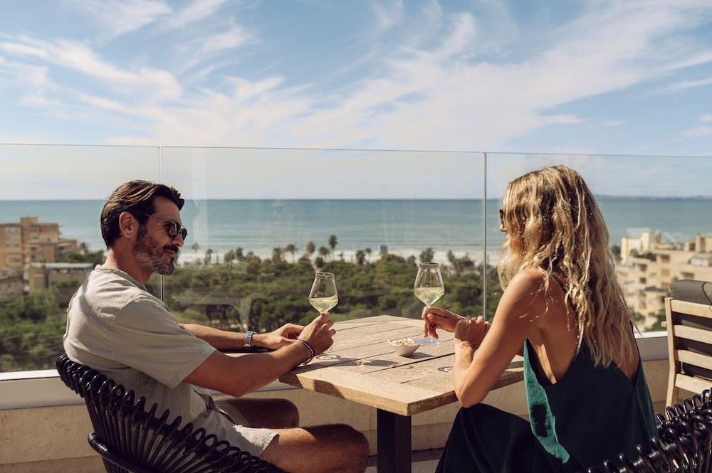 Zwei Personen trinken Weißwein auf einem sonnigen Balkon mit Blick auf Meer und Stadt. Iberostar Selection Llaut Palma – Nur für Erwachsene
