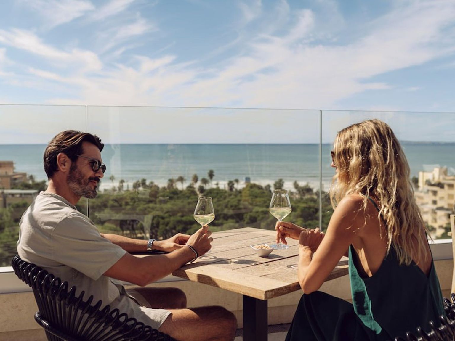 Zwei Personen trinken Weißwein auf einem sonnigen Balkon mit Blick auf Meer und Stadt. Iberostar Selection Llaut Palma – Nur für Erwachsene