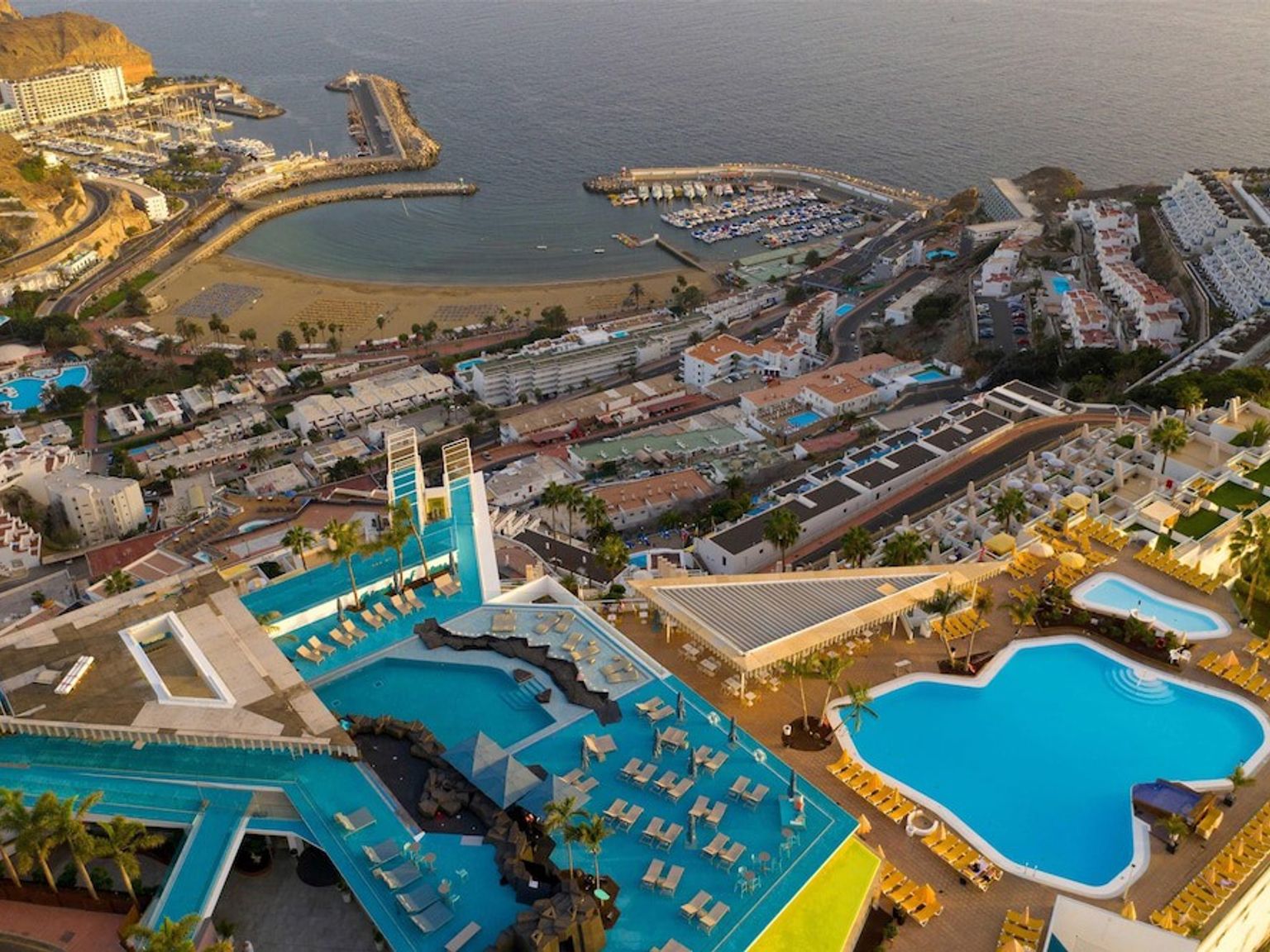 Flygfoto av en kustort med flera swimmingpooler, en marina och en strand.