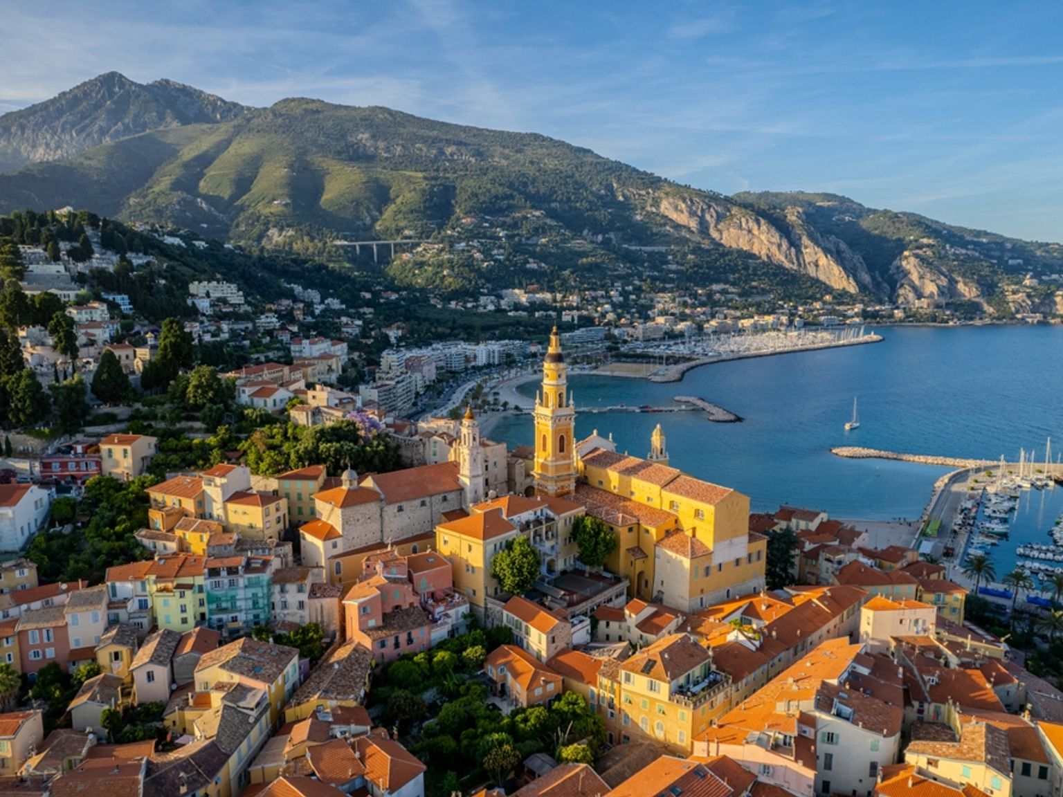 Ilmakuva värikkäästä rannikkokaupungista, jossa on näkyvä keltainen kirkko, venesatama, ranta ja vuoria sinisen taivaan alla. Menton, Ranska