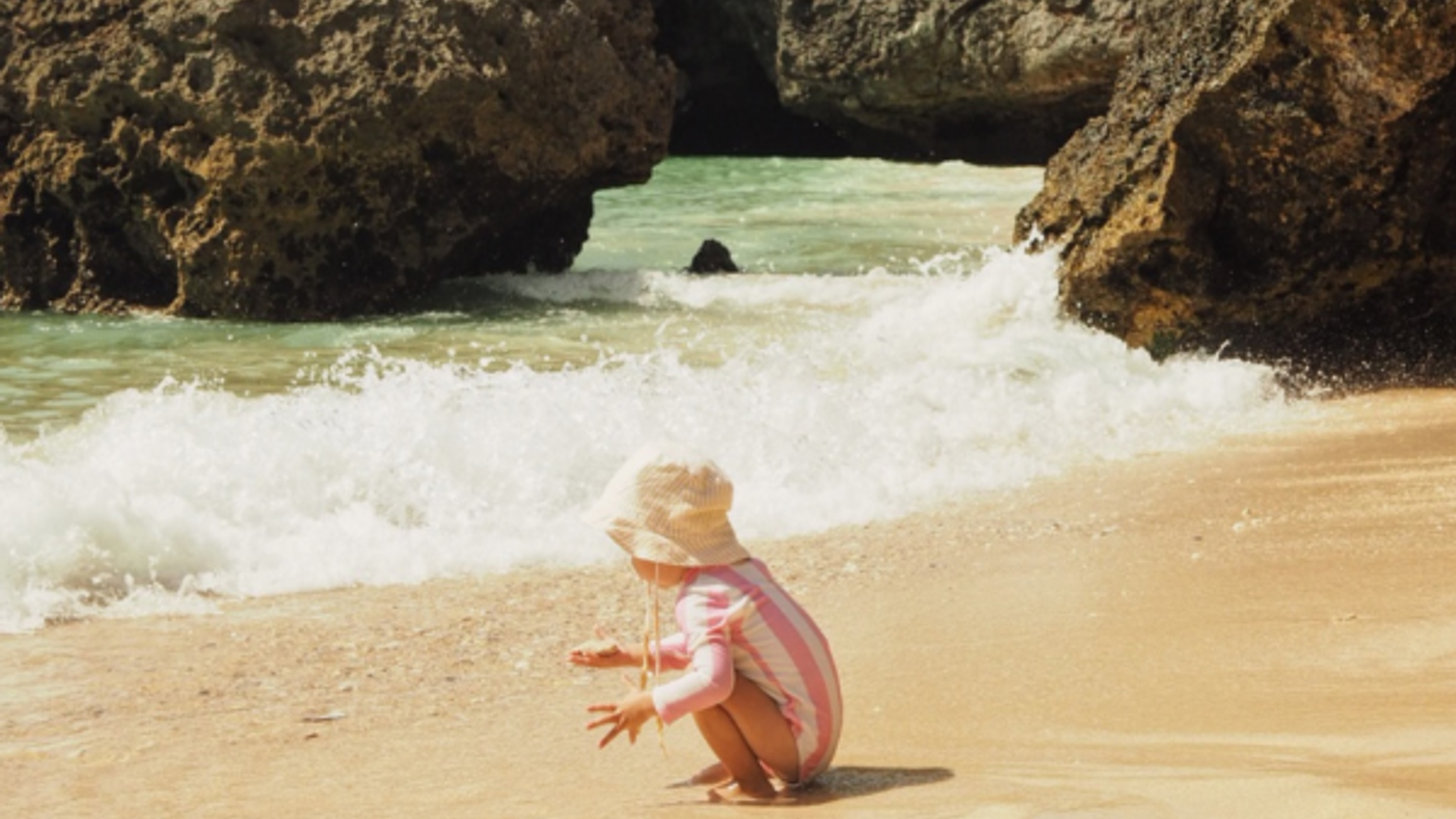 Ett barn i hatt och randig baddräkt leker vid vågorna på en sandstrand med stenar i bakgrunden.