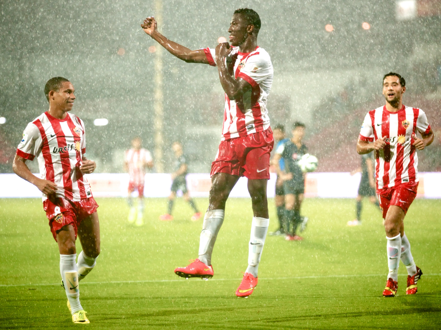 Drei Fußballspieler in rot-weißen Trikots auf einem Spielfeld im strömenden Regen, wobei einer in die Luft springt und jubelt.