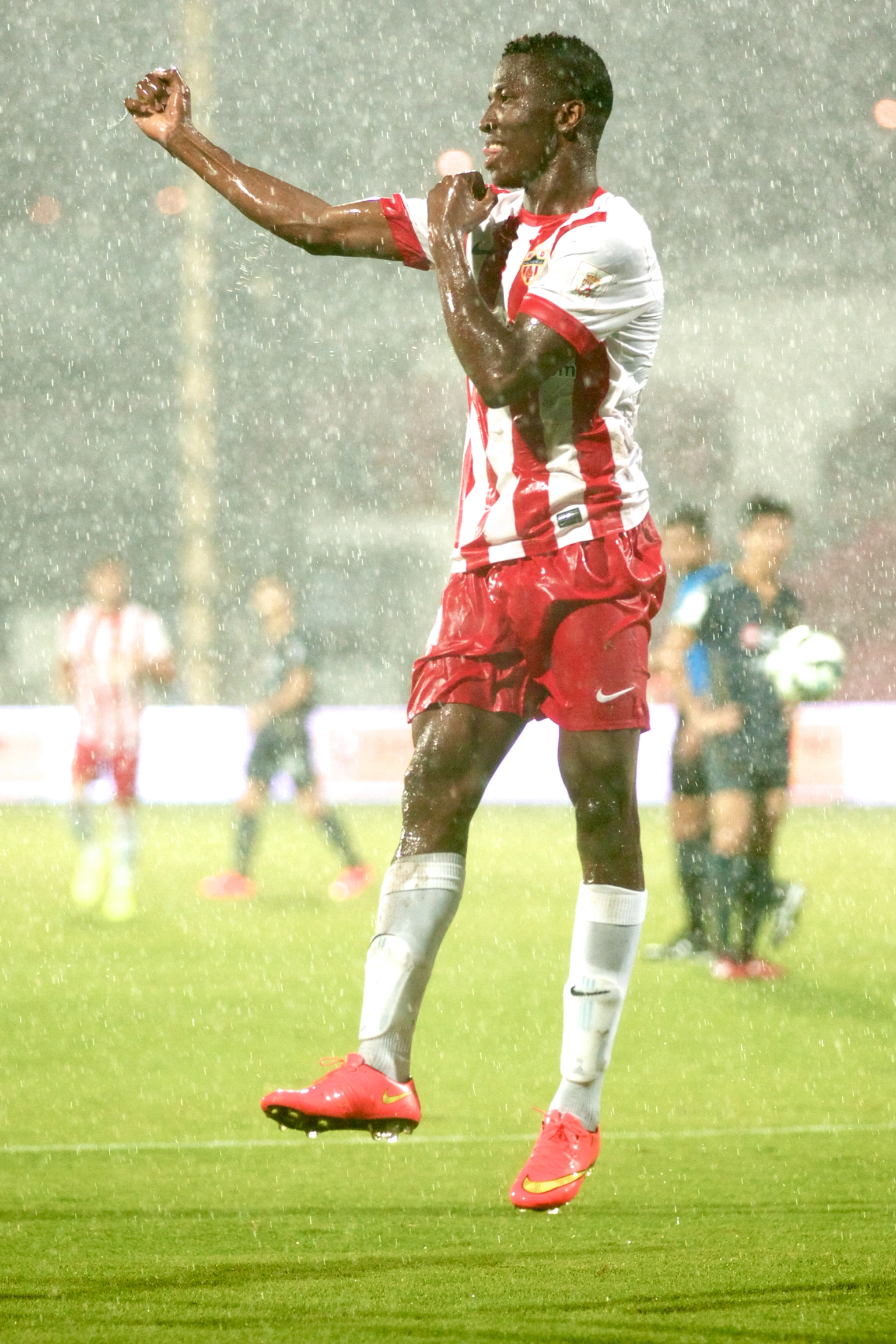 Drei Fußballspieler in rot-weißen Trikots auf einem Spielfeld im strömenden Regen, wobei einer in die Luft springt und jubelt.