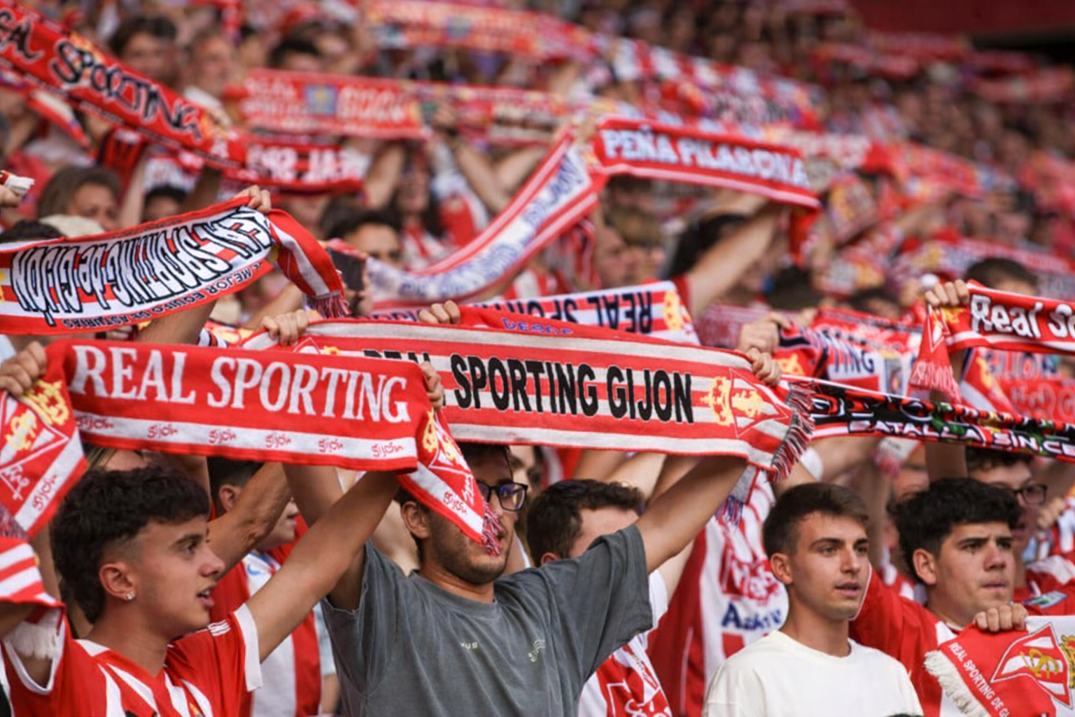 A crowd of football fans are holding up red and white Real Sporting Gijón scarves.