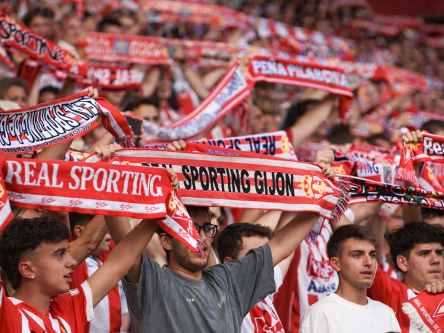 A crowd of football fans are holding up red and white Real Sporting Gijón scarves.