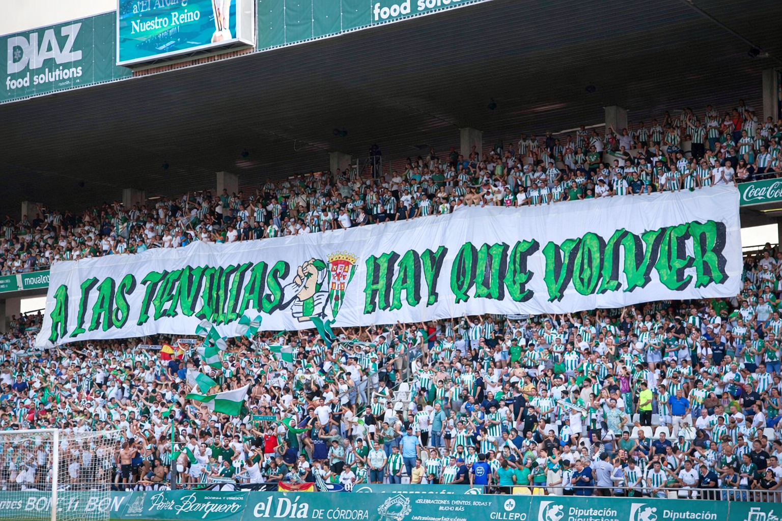 A large crowd of soccer fans in green and white shirts hold up a white banner with green text that reads "A LAS TENDILLAS HAY QUE VOLVER" in a stadium.