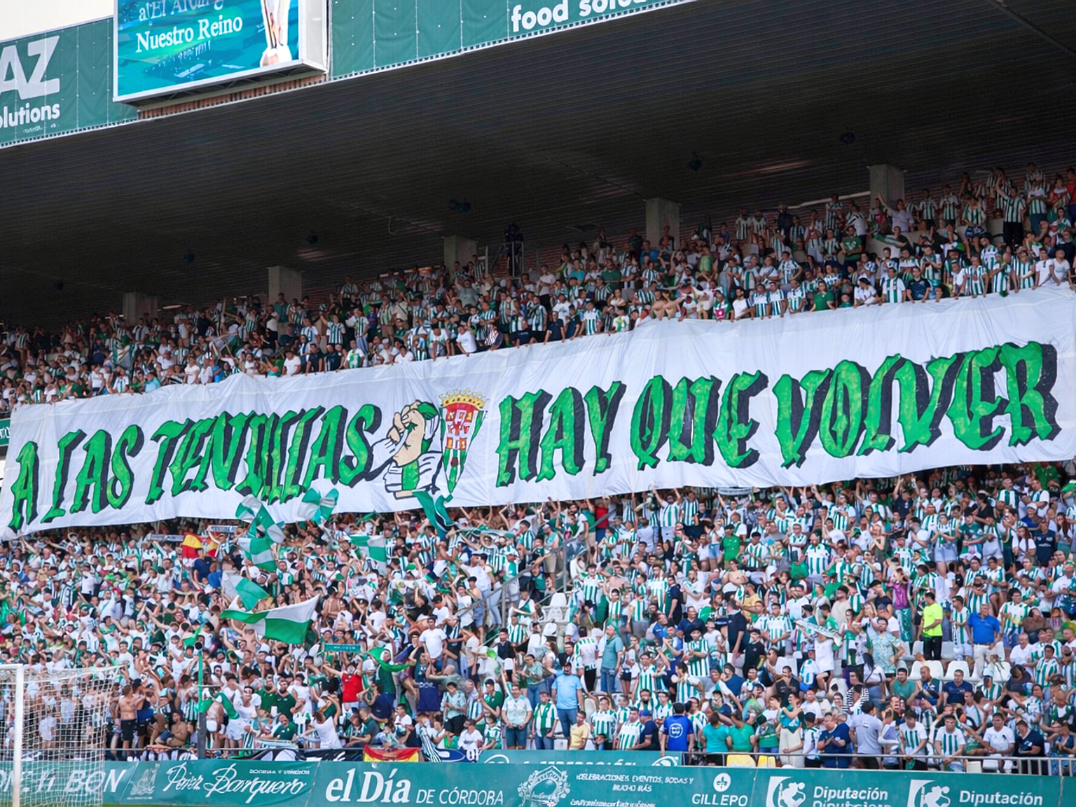 A large crowd of soccer fans in green and white shirts hold up a white banner with green text that reads "A LAS TENDILLAS HAY QUE VOLVER" in a stadium.