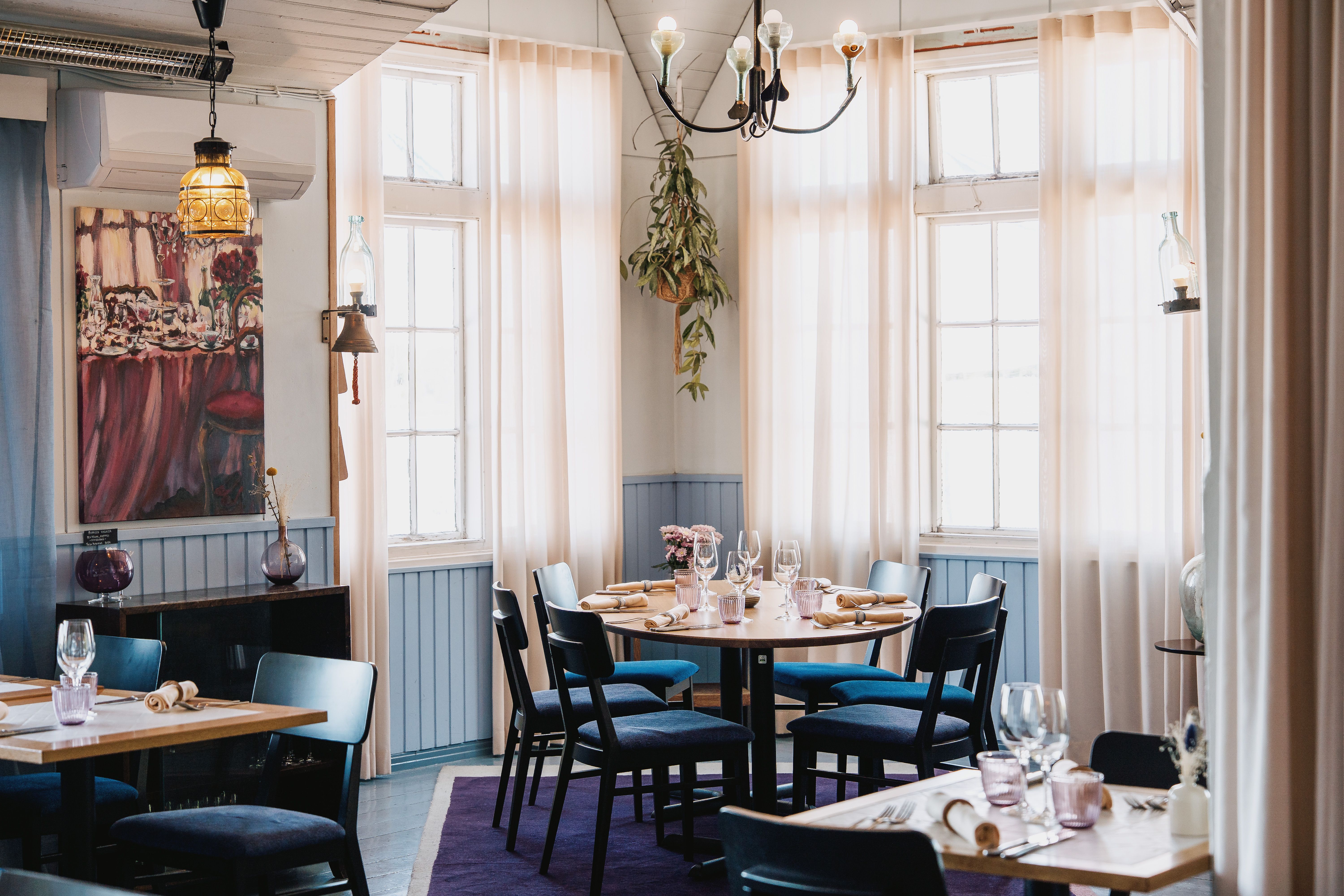 En lys, elegant restaurantspisestue med lyseblå brystpaneler, store vinduer med lette gardiner og flere borde opstillet med mørke stole.