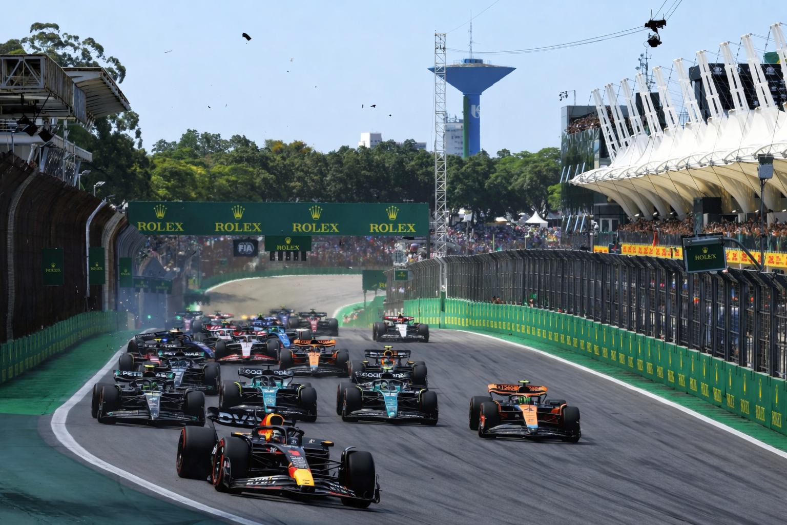 Numerous Formula 1 cars race on a track, passing grandstands filled with spectators.